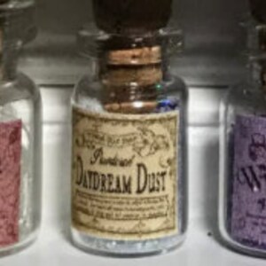 Puede incluir: Tres pequeñas botellas de vidrio transparente con tapones de corcho. La botella central tiene una etiqueta que dice "Daydream Dust" en una fuente antigua. Las etiquetas de las botellas son de varios colores, incluyendo rosa y morado.