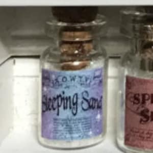 Könnte beinhalten: Kleine, klare Glasflasche mit Korken und Etikett mit der Aufschrift „Sleeping Sand“. Das Etikett ist hellblau und lila mit weißem Text und Sternillustrationen. Die Flasche ist mit einer weißen, körnigen Substanz gefüllt.
