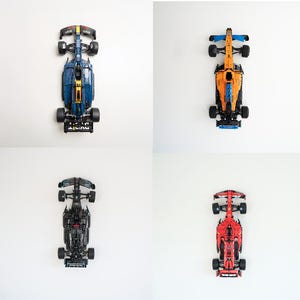 May include: Four toy race cars in blue, orange, black, and red are displayed against a white background. Each car is a detailed model, showcasing the design of a Formula 1 race car. The cars are positioned to show their top view.