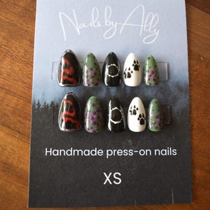 May include: A set of handmade press-on nails in XS size. The nails feature various designs, including red swirls on black, green with purple dots, black with white accents, and white with black paw prints. The card reads "Nails by Ally".