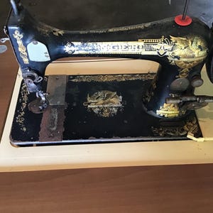 Puede incluir: Máquina de coser Singer antigua, negra con detalles dorados. La máquina presenta diseños ornamentados y una base color crema. El logotipo de Singer es visible. Una herramienta de costura vintage para proyectos de costura y textiles.