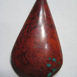Puede incluir: Una gema en forma de lágrima con una superficie pulida. La piedra es principalmente de color rojo intenso, salpicada de negro y acentuada con parches de turquesa. La gema tiene una base lisa y redondeada y termina en punta.