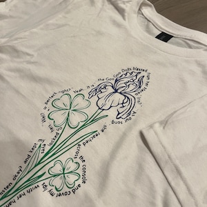 以下が含まれることがあります： 緑と青の花柄が特徴の白いTシャツ。デザインには、青いアヤメの花と四つ葉のクローバーが含まれています。「Yeah... It is」や「As our song」などのテキストが含まれています。