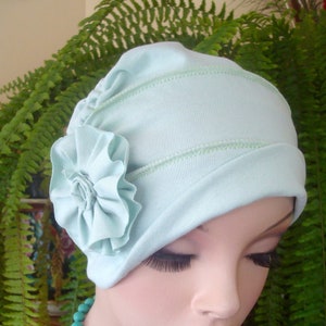 May include: A light blue cloche hat with a ruffled flower accent. The hat has a stitched design and is displayed on a mannequin. A turquoise beaded necklace is also visible.