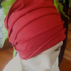 Op de afbeelding: Een rood stoffen hoofdband met meerdere plooien en naden. Het hoofdband wordt gedragen op het hoofd van een mannequin.
