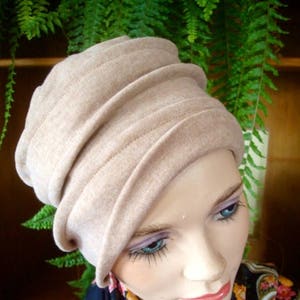 Puede incluir: Un gorro de punto beige con un diseño plegado. El gorro lo lleva una persona con una bufanda azul oscuro con estampado floral.