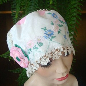 Può includere: Un berretto di lino bianco con fiori ricamati in rosa, blu e verde. Il berretto ha un bordo di pizzo e un fiore di stoffa rosa.