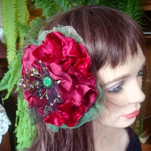 Puede incluir: Una pinza para el pelo de terciopelo rojo con adornos verdes y marrones. La flor es grande y tiene una textura rizada. La pinza est&aacute; unida a una malla verde y marr&oacute;n transparente.