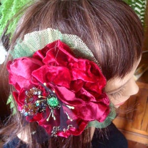 Puede incluir: Un clip de pelo de terciopelo rojo con una joya verde y cuentas marrones. La flor est&aacute; unida a una malla negra y verde transparente.
