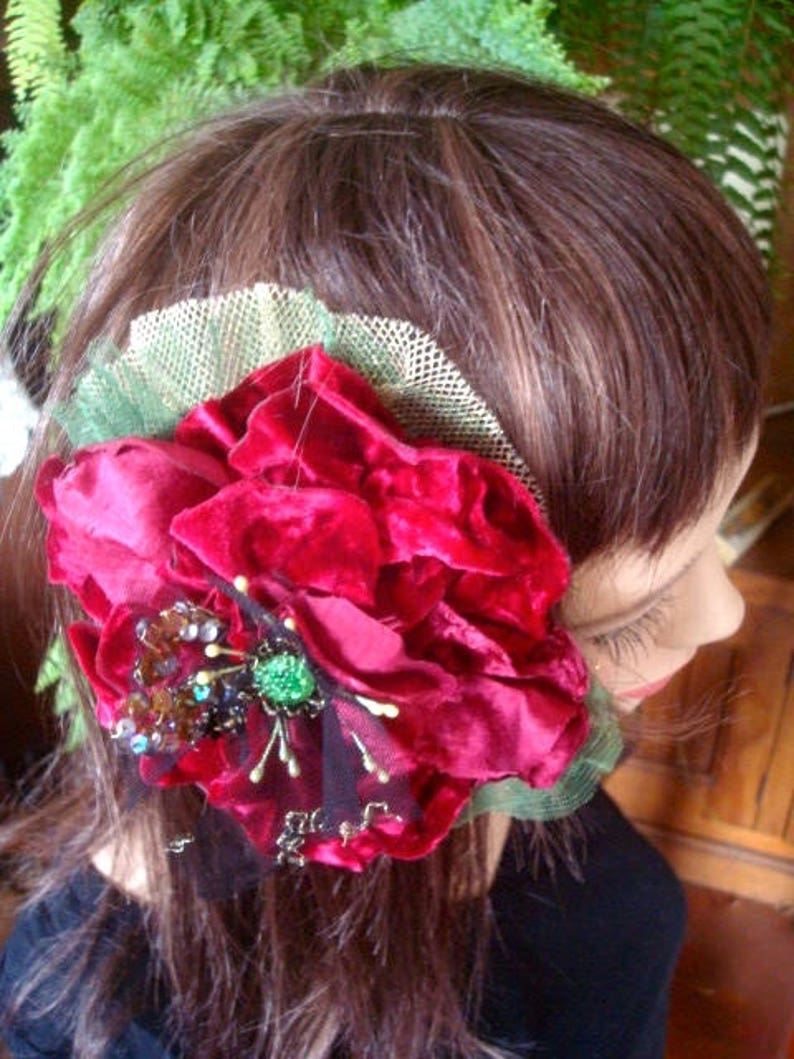 Puede incluir: Un clip de pelo de terciopelo rojo con una joya verde y cuentas marrones. La flor est&aacute; unida a una malla negra y verde transparente.