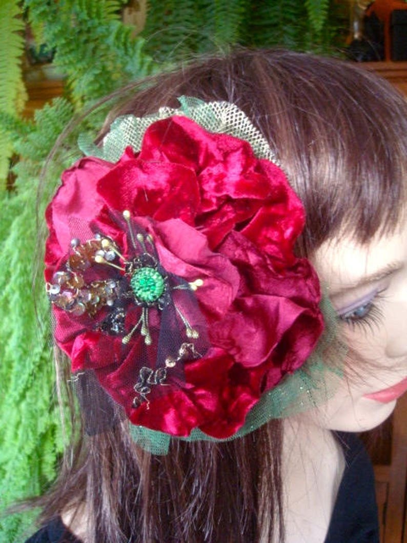 Puede incluir: Una pinza para el pelo de terciopelo rojo con adornos verdes y dorados. La flor est&aacute; en capas y tiene una base de tul.