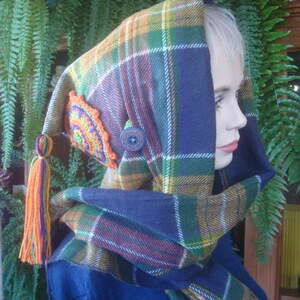 May include: A hooded scarf with a plaid pattern in shades of green, blue, orange, and white. The scarf has a crocheted flower detail and a button.