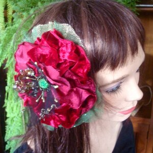 Puede incluir: Una pinza para el pelo de terciopelo rojo con una red verde y adornos dorados y negros. La pinza se lleva en el pelo casta&ntilde;o de una mujer.
