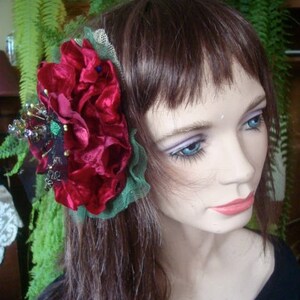 Puede incluir: Una pinza para el pelo de terciopelo rojo con hojas verdes y un adorno de cuentas plateadas y negras. La pinza se lleva en el pelo casta&ntilde;o de una mujer.