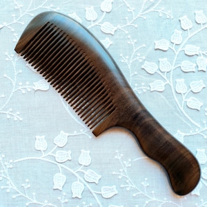 May include: A dark brown wooden comb with a curved handle and fine teeth. The comb is made of a smooth, polished wood and is designed for hair grooming. The comb is set against a white floral patterned background.
