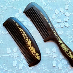 May include: Two dark brown wooden combs with fine teeth. The combs are decorated with gold floral patterns along the top edge. One comb has a handle, and the other is smaller. The combs are displayed on a light blue surface.