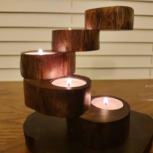 May include: A wooden candle holder with four candle holders in a cascading design. The candle holder is made of dark brown wood and has a natural finish. The candles are lit and glowing.