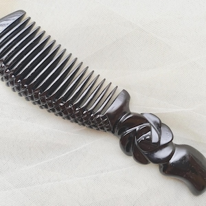 May include: A dark brown, polished hair comb featuring a curved row of teeth and a handle carved into a layered, rounded floral shape. The comb has a textured, cross-hatched pattern along the spine, providing a detailed grip and aesthetic.