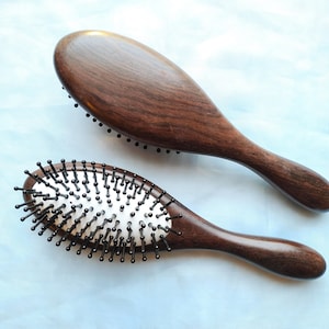 May include: Two wooden hairbrushes with dark brown handles and bristles. One brush is larger and has a smooth wooden surface, while the other has a silver base with black bristles. The brushes are oval-shaped.