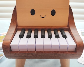May include: A wooden toy piano with a smiling face on the backboard. The piano has white and black keys, and is supported by four small wooden legs. The wood has a warm, reddish-brown tone.
