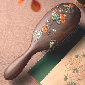 May include: A wooden hairbrush with a dark brown finish. The brush has an oval head and a long handle. The head is decorated with a branch of orange persimmons and green leaves. The bristles are black.