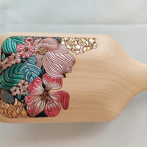 May include: Wooden hairbrush with a floral design. The brush features a light wood finish and a detailed carving of pink, teal, and cream-colored flowers and leaves on the top surface. The handle is integrated into the design.