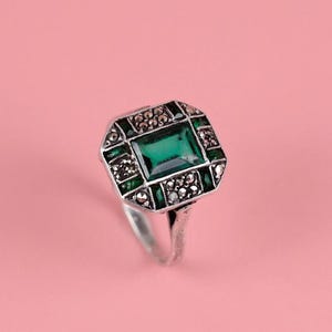 Könnte beinhalten: Ein silberfarbener Ring mit einem rechteckigen grünen Edelstein in der Mitte, der von einer Umrandung aus kleinen, dunklen, metallischen Akzenten umgeben ist. Die Fassung hat eine achteckige Form mit feinen Details am Rand.