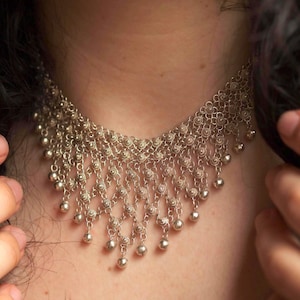 Könnte beinhalten: Silberfarbene Halskette mit filigranem, aufwendigem Design. Die Halskette weist eine Reihe miteinander verbundener Blumenmuster und hängende kugelförmige Perlen auf. Der Schmuck ist zum Tragen um den Hals konzipiert.