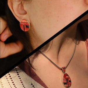 Könnte beinhalten: Eine silberne Halskette mit Anhänger und passende Ohrstecker. Der Schmuck weist ein geometrisches Design in Rot, Schwarz und Weiß auf. Der Anhänger ist tropfenförmig und die Ohrringe sind rund. Der Schmuck wird von einer Person getragen.