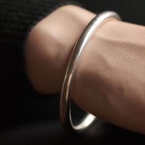 Könnte beinhalten: Ein silberfarbenes, abgerundetes Armreif-Armband. Das Armband wird an einem Handgelenk getragen, die Haut ist sichtbar. Das Armband hat eine glatte, reflektierende Oberfläche. Der Hintergrund ist dunkel, was das helle Aussehen des Armbands betont. Der Schmuck ist schlicht und elegant.