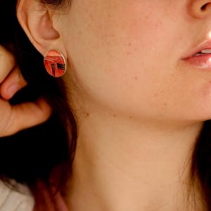 Könnte beinhalten: Ohrstecker in ovaler Form mit silbernem Rand. Der Ohrring hat ein rotes und schwarzes geometrisches Design. Der Ohrring wird am Ohr einer Person getragen.
