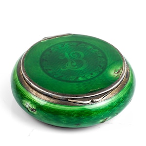 Puede incluir: Pequeña caja redonda esmaltada en verde con borde plateado. La tapa presenta una letra "B" estilizada en un tono verde más claro. La caja tiene una forma ligeramente abovedada y un acabado brillante.