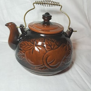 Pode incluir: Bule de cerâmica castanho com tampa e pega de metal. O bule apresenta desenhos de frutas em relevo e um bico. A pega é feita de metal enrolado. O bule é de cor castanho escuro com detalhes em castanho claro.