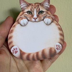 May include: A die-cut sticky note shaped like a round, orange tabby cat with white paws and a white belly. The cat has its front paws resting on the top of its large, circular, blank white centre area, which is intended for writing notes.