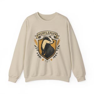 May include: A cream-colored sweatshirt featuring a Hufflepuff crest. The crest includes a badger, the house name, and decorative elements in yellow, black, and brown. The sweatshirt has long sleeves and a crew neck.