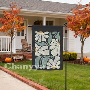 May include: A black garden flag featuring a floral design with white and light green flowers and leaves. The flag is displayed on a black metal stand in a front yard with a house and fall foliage in the background. The word "Chanyya" is visible.