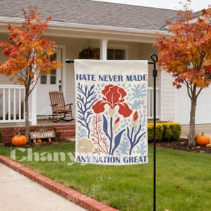May include: A cream-coloured garden flag featuring the words "HATE NEVER MADE ANY NATION GREAT" and a floral design in red, blue, and beige. The flag is displayed outdoors in front of a house with autumn leaves.