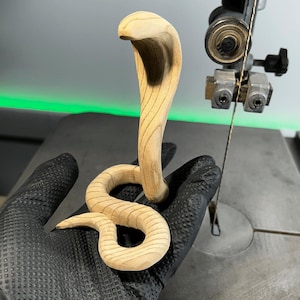 May include: A wooden cobra sculpture, carved with intricate details, is held in a gloved hand. The sculpture is light brown and has a smooth, polished finish.