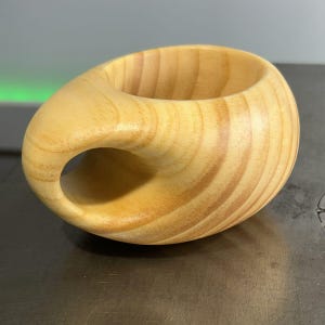 Op de afbeelding: Een houten beker met een unieke, gebogen handgreep. De beker is gemaakt van lichtbruin hout met zichtbare houtnerf.