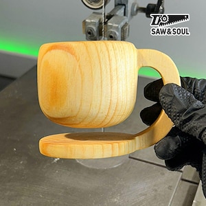 May include: A wooden cup with a handle being cut out on a band saw. The cup is made of light brown wood and has a natural finish. The saw is cutting through the wood, leaving a smooth, clean cut.