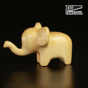 May include: A small, light-coloured wooden elephant figurine with a trunk, ears, and four legs. The figurine is carved from a single piece of wood and has a smooth, polished finish. The logo "SAW&SOUL" is in the upper right corner.