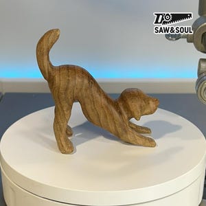 May include: A wooden sculpture of a dog in a stretching pose. The dog is carved from light brown wood, with visible grain patterns. The sculpture is on a white surface, with the "SAW&SOUL" logo in the top right corner.