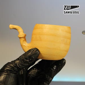 May include: A light-coloured wooden pipe with a curved stem and a rounded bowl. The pipe is made of wood, showing the natural grain. The logo "SAW&SOUL" is visible in the upper right corner.