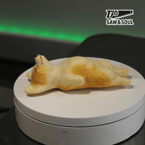 Könnte beinhalten: Holzfigur eines Corgi, der auf dem Rücken liegt und die Augen geschlossen hat. Der Hund hat helles Fell mit orange-braunen Flecken. Die Figur steht auf einer weißen, kreisförmigen Plattform. Das Logo "SAW & SOUL" ist oben rechts zu sehen.