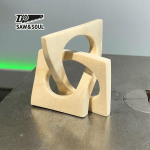 Op de afbeelding: Een houten sculptuur gemaakt van drie in elkaar grijpende stukken, elk met een cirkelvormige uitsparing. De sculptuur is lichtbruin van kleur en heeft een gladde, gepolijste afwerking.