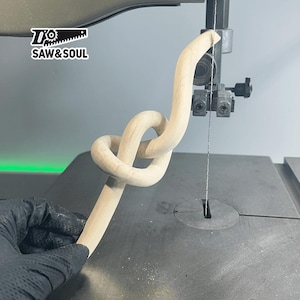 Pode incluir: Um nó de madeira está sendo cortado em uma serra de fita. O nó está sendo segurado por uma mão enluvada. A serra está cortando através do nó, deixando um corte liso e limpo. O logotipo "Saw & Soul" é visível no fundo.