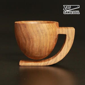 Puede incluir: Taza de café de madera hecha a mano con un asa y base curvadas únicas. La taza es de madera marrón claro, mostrando la veta natural. El logotipo "SAW&SOUL" es visible. El diseño de la taza es moderno.