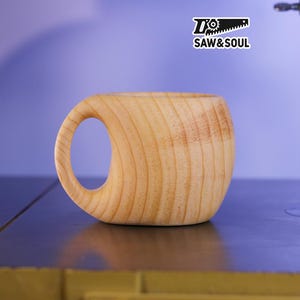 May include: A handcrafted wooden mug with a large, rounded handle. The mug is made from light-colored wood, showcasing the natural grain. The text "SAW&SOUL" is printed on a white sticker in the upper right corner.