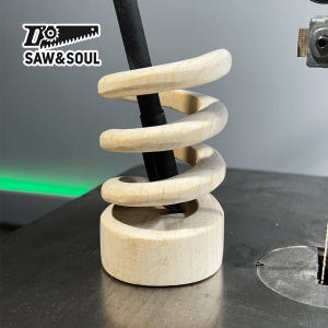 Può includere: Un portapenne in legno a forma di spirale, con una penna nera inserita. Il portapenne è in legno chiaro e ha una finitura liscia. Il portapenne è su una superficie nera. Il testo "SAW & SOUL" è visibile sullo sfondo.