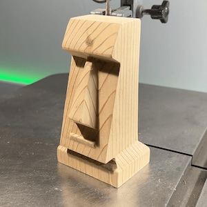 Könnte beinhalten: Eine Holzschnitzerei eines Gesichts, möglicherweise ein Tiki, die auf einer Bandsäge geschnitten wird. Die Schnitzerei ist aus hellem braunem Holz und hat ein einfaches Design.
